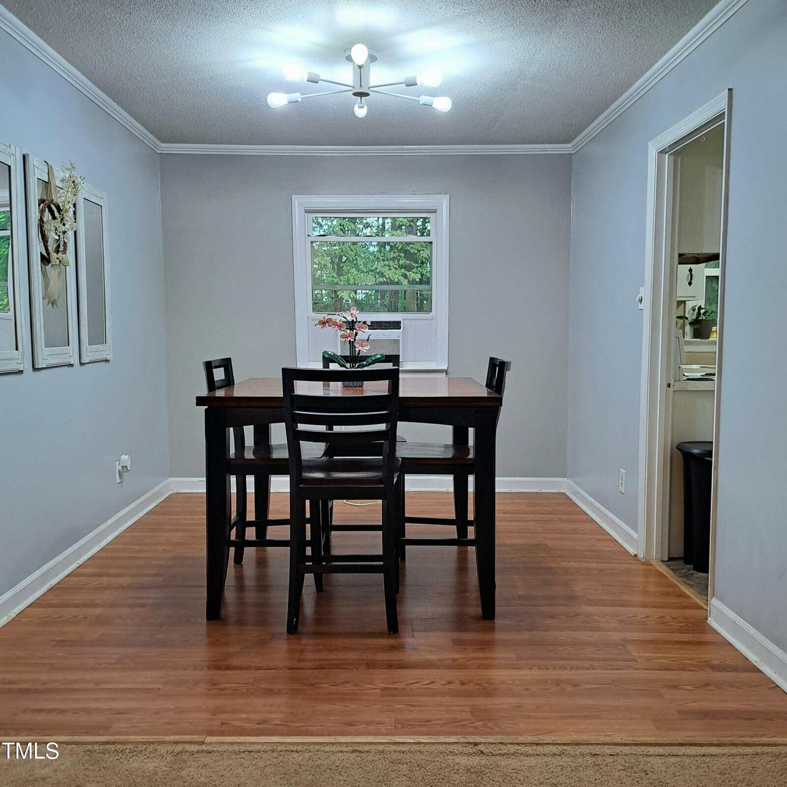 A dining room with a dark wooden table and six matching chairs on a hardwood floor. A modern chandelier hangs overhead. There are three framed artworks on the left wall and a window displaying green foliage at the far end. A doorway leads to another room.