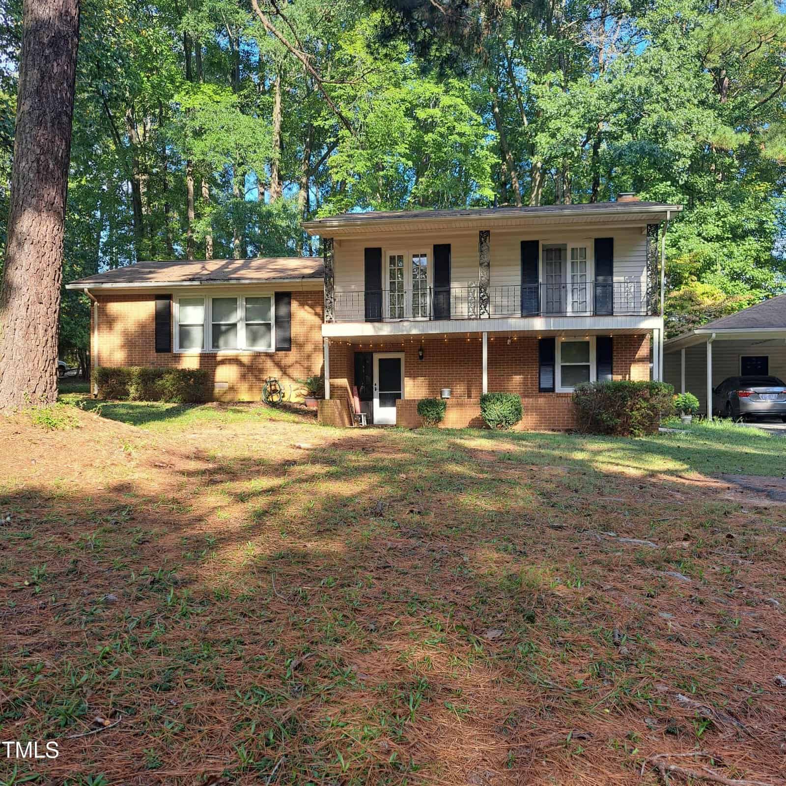 A two-story brick house with a balcony on the second floor, black shutters, and a front porch. The yard is covered with pine needles and surrounded by trees. A driveway on the right leads to a carport.