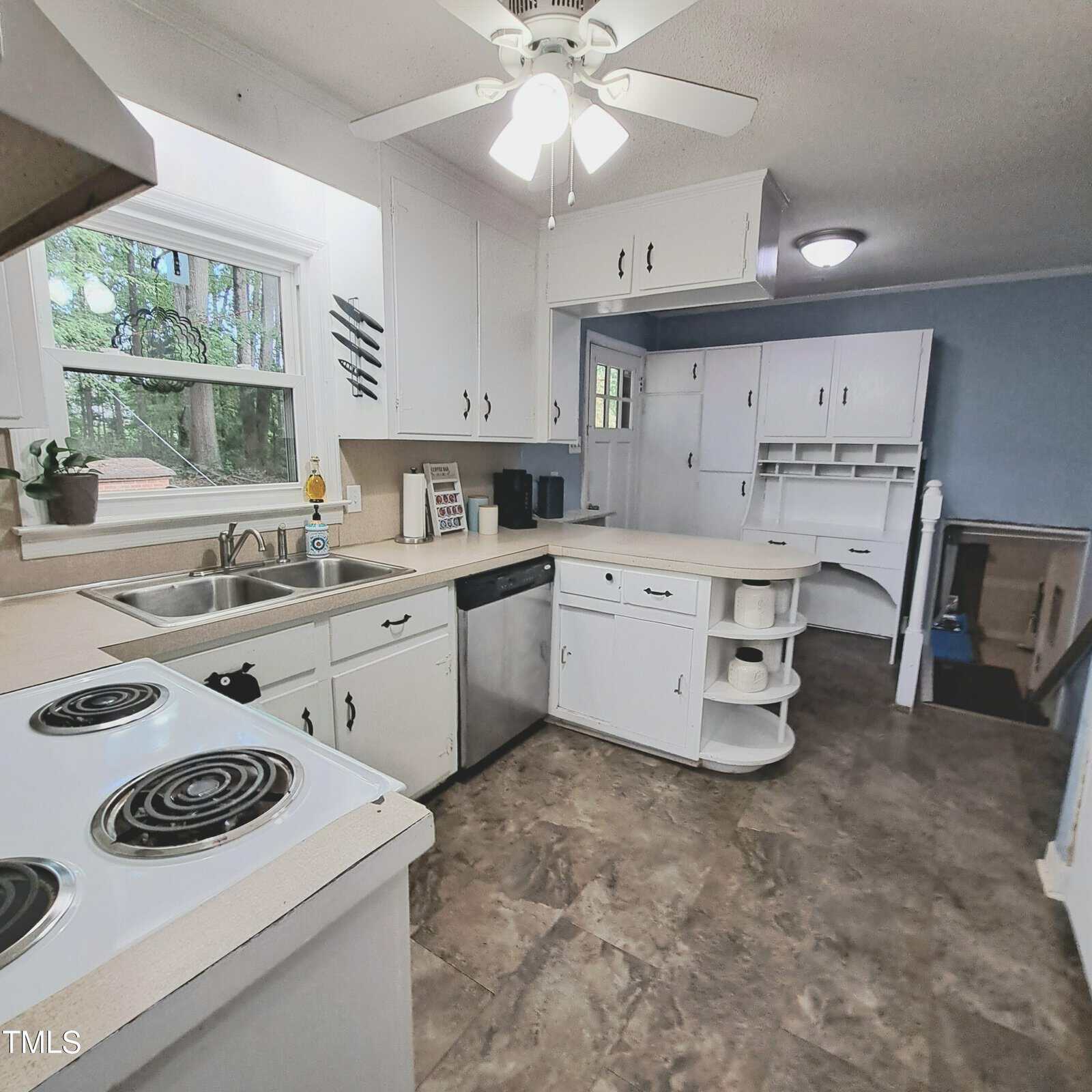 A kitchen with white cabinets and countertops, featuring a stove, sink, and dishwasher. A ceiling fan with lights is above, and the floor has patterned tiles. A window above the sink shows trees outside. Shelving and a small staircase are visible.