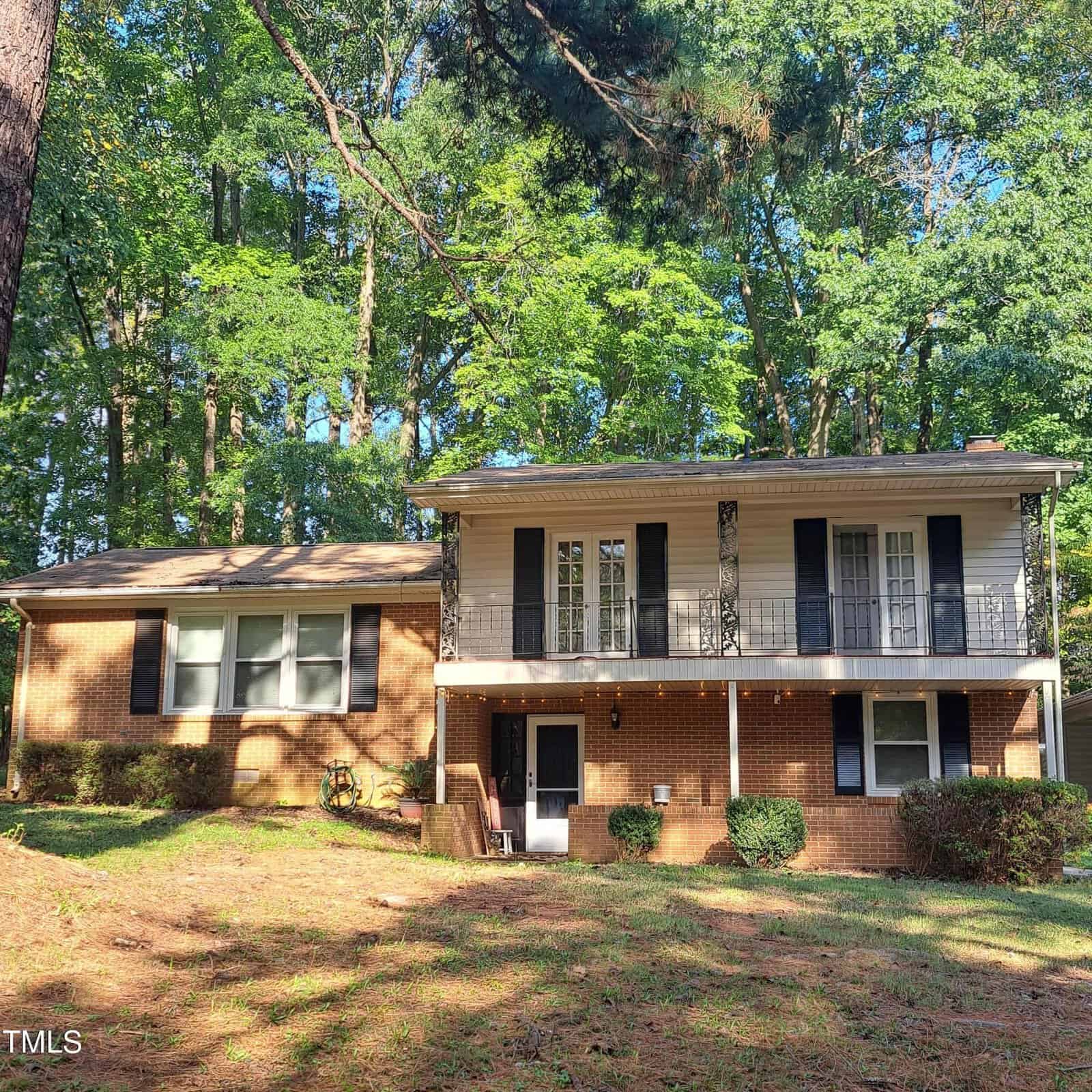Nestled at 123 Maluli Drive in Oxford, this charming two-story brick house boasts white accents and is surrounded by trees. The upper level features a balcony with black railings, while the shaded yard below is blanketed with grass and pine needles, enhancing its serene appeal.