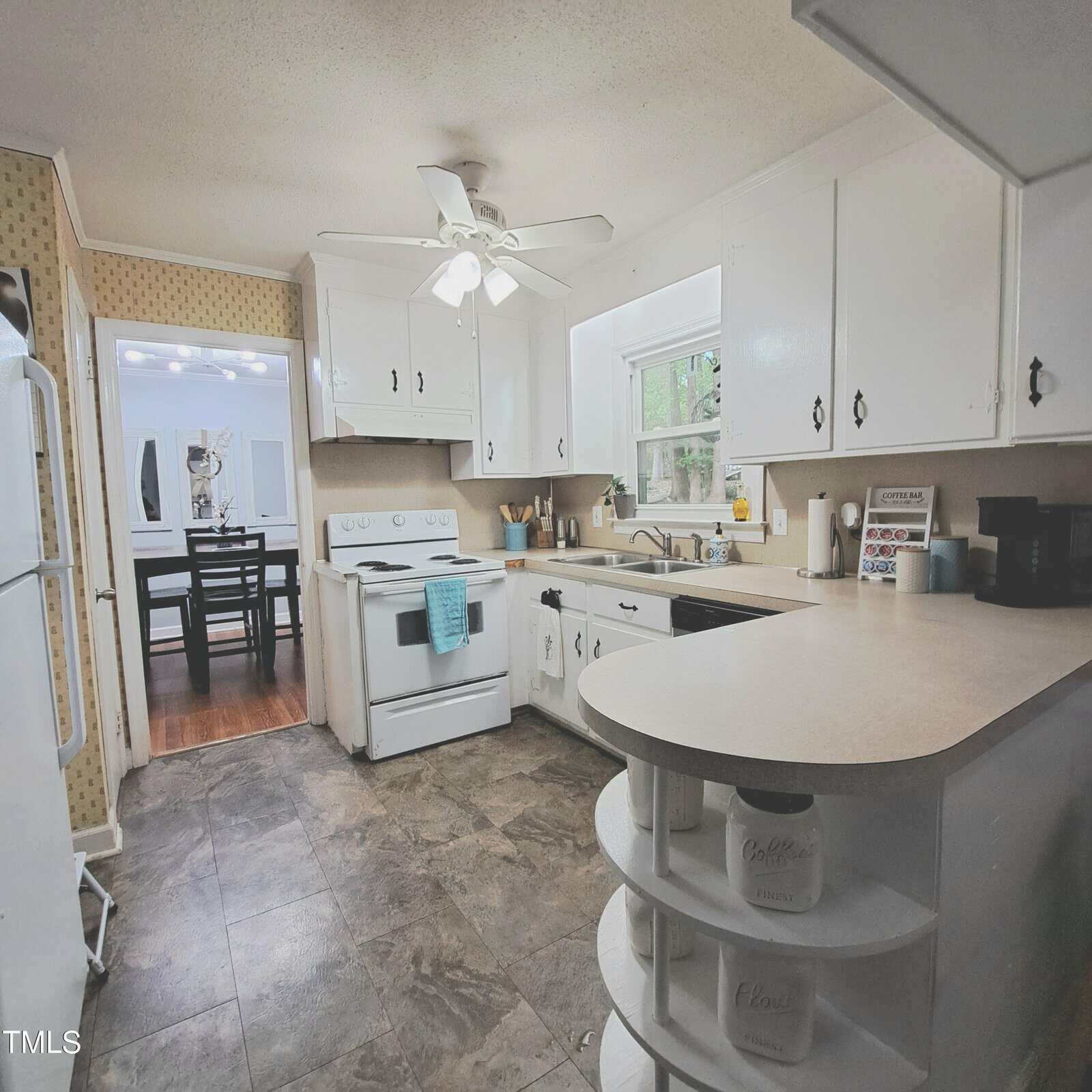 A kitchen with white cabinets and countertops, featuring a white stove, refrigerator, and a window above the sink. A ceiling fan is installed overhead. The floor has dark tiles, and a doorway leads to a dining area with a table and chairs.