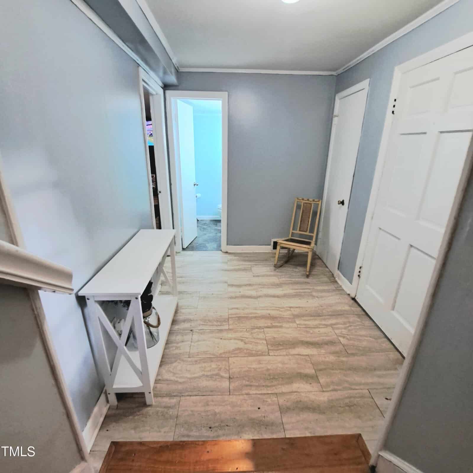 A hallway with light blue walls, beige tiled flooring, and white trim. It features a white console table on the left and a wooden chair in the corner near the doorways. There are three white doors and one open doorway leading to another room.