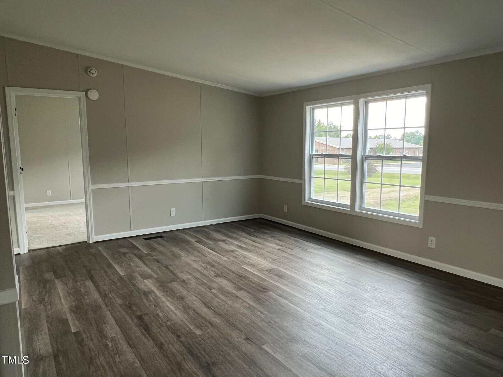 A vacant living room with light gray walls, white trim, and dark wood laminate flooring. Large windows on the right side let in natural light, and an open doorway on the left leads to another room. A smoke detector is visible on the wall.