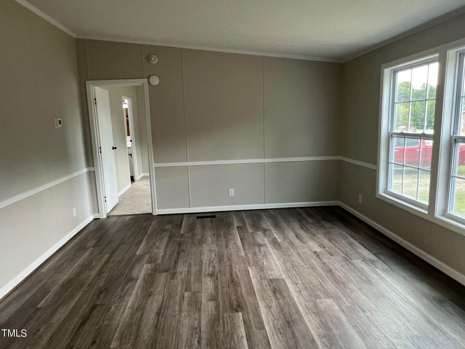 A bright, empty room with light beige walls, white trim, and wood-look laminate flooring. Large windows on the right allow natural light to enter. An open doorway on the left leads to another room, and another door is visible in the background.