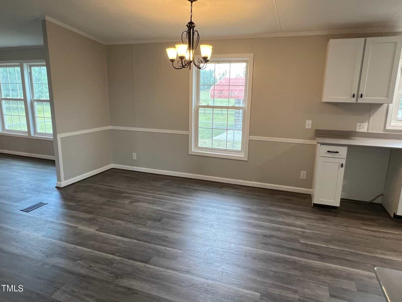 A bright, empty room with light gray walls, wood-look flooring, and a central window with a view of the outside. It features a white trim and baseboards, a light fixture hanging from the ceiling, and a glimpse of adjacent rooms and the kitchen area.