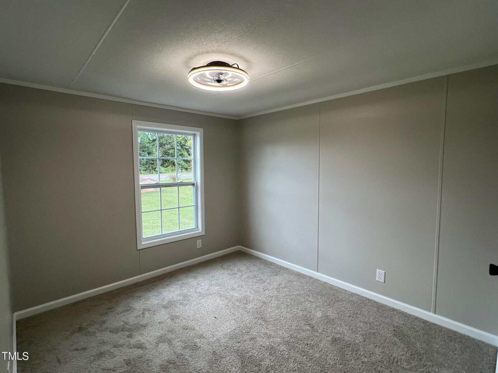 An empty room with beige walls and carpet, featuring a ceiling light fixture. A single rectangular window provides a view of a grassy area outside, allowing natural light to enter the room.