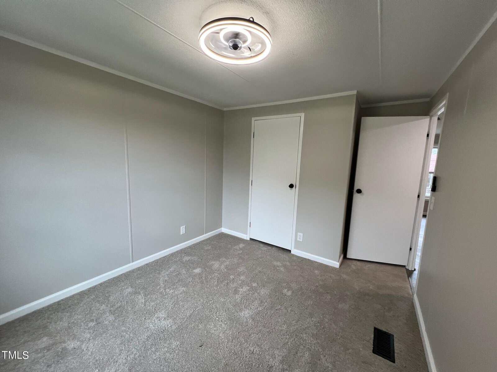 A small, empty room with beige carpet, light gray walls, and a white ceiling. It has a round ceiling light fixture, a closed white door, and an ajar door leading to another space. A small floor vent is visible in the corner.