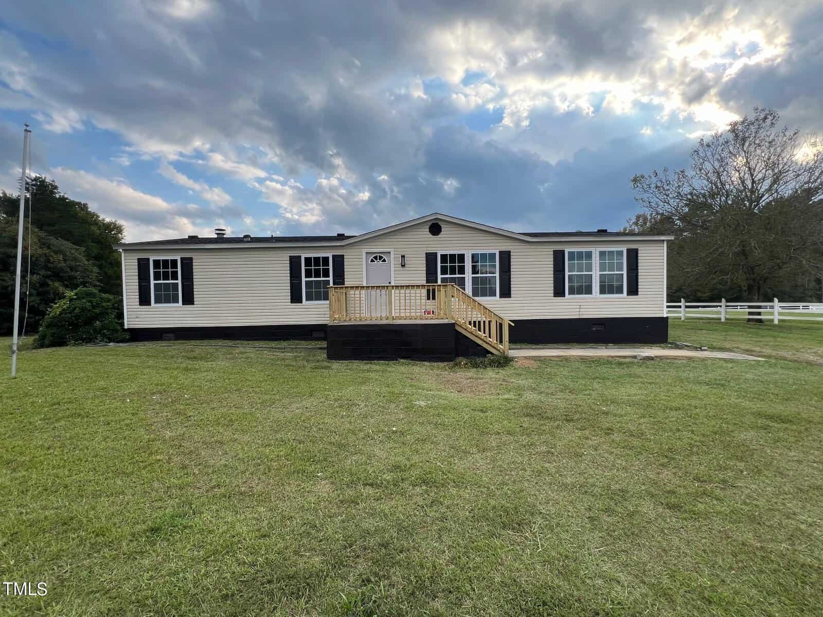 A single-story manufactured home with beige siding and a black foundation sits on a grassy lawn at 805 Morgan Road in Henderson. It features a small wooden porch with steps leading to the front door. The sky above is partly cloudy, and there's a tree and a white fence in the background.