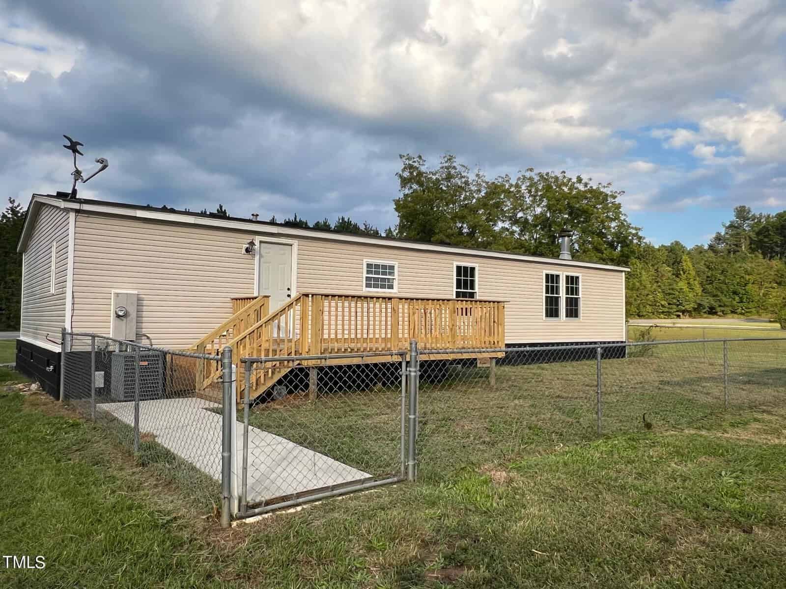 A single-story beige mobile home with a wooden front porch sits on a grassy lawn, surrounded by a chain-link fence. The home is set against a backdrop of trees and a partly cloudy sky. An antenna is visible on the roof.