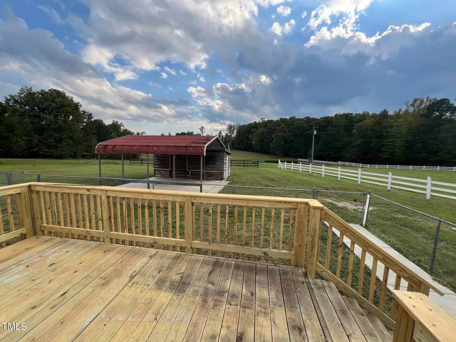 A wooden deck overlooks a spacious, grassy yard with a small red-roofed shed. The area is fenced with white fencing, and the background features trees and a partly cloudy sky.