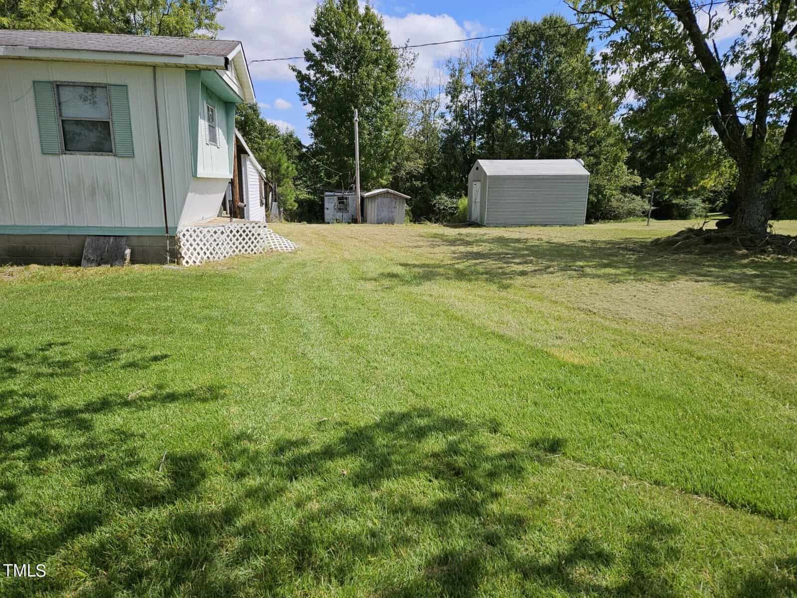 A modest mobile home with light green siding sits on the left side of a large, well-manicured grassy yard. In the background, there are two small outbuildings, and numerous trees surrounding the property. Power lines are visible overhead. The sky is partly cloudy.