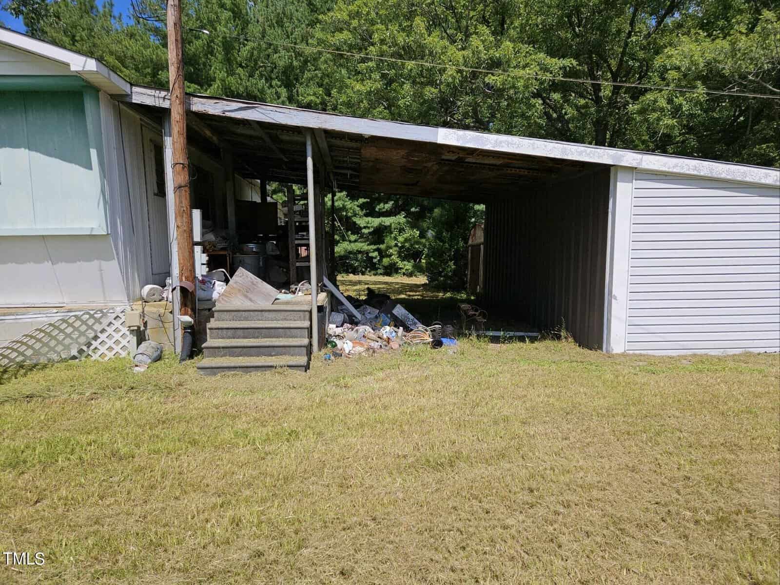 A dilapidated, partially enclosed carport is attached to a mobile home. The area underneath is cluttered with trash and various items. The roof of the carport appears to be in poor condition. The surrounding yard is grass-covered, and trees are visible in the background.