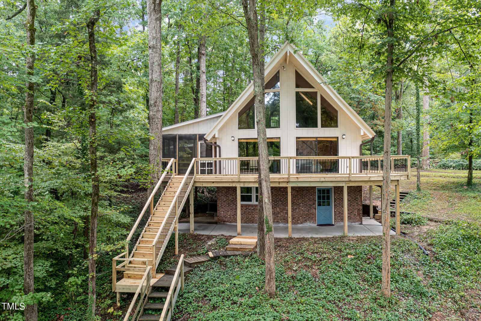 A two-story A-frame cabin at 148 Kerr Lake Road features a mix of wood and brick exterior, nestled in a forested area of Henderson. Large triangular windows, a wraparound deck, and an elevated staircase leading to the main entrance harmonize with the lush green trees and foliage surrounding the property.