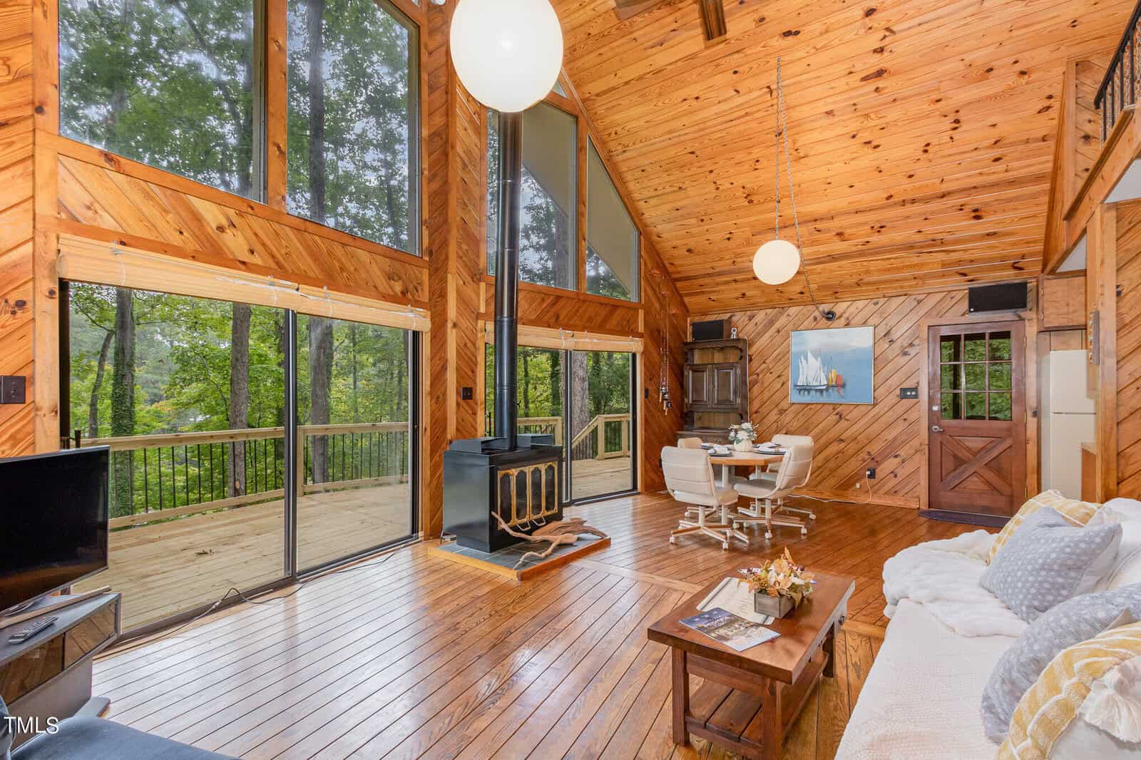 A cozy wooden cabin interior with tall windows and a high ceiling. It features a living area with a couch and coffee table, a wood-burning stove, and a dining area. Sliding glass doors open to a wooden deck, offering a view of the lush forest outside.