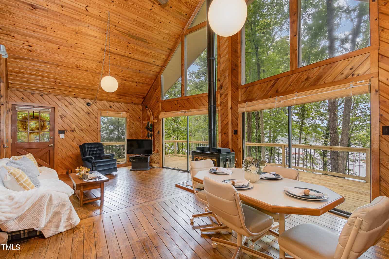 A cozy wooden cabin interior with high ceilings, large windows, and a spacious open layout. The room features a comfortable seating area with a sofa and armchair, a TV, and a dining table with chairs. The windows offer a view of lush greenery and a glimpse of a lake.