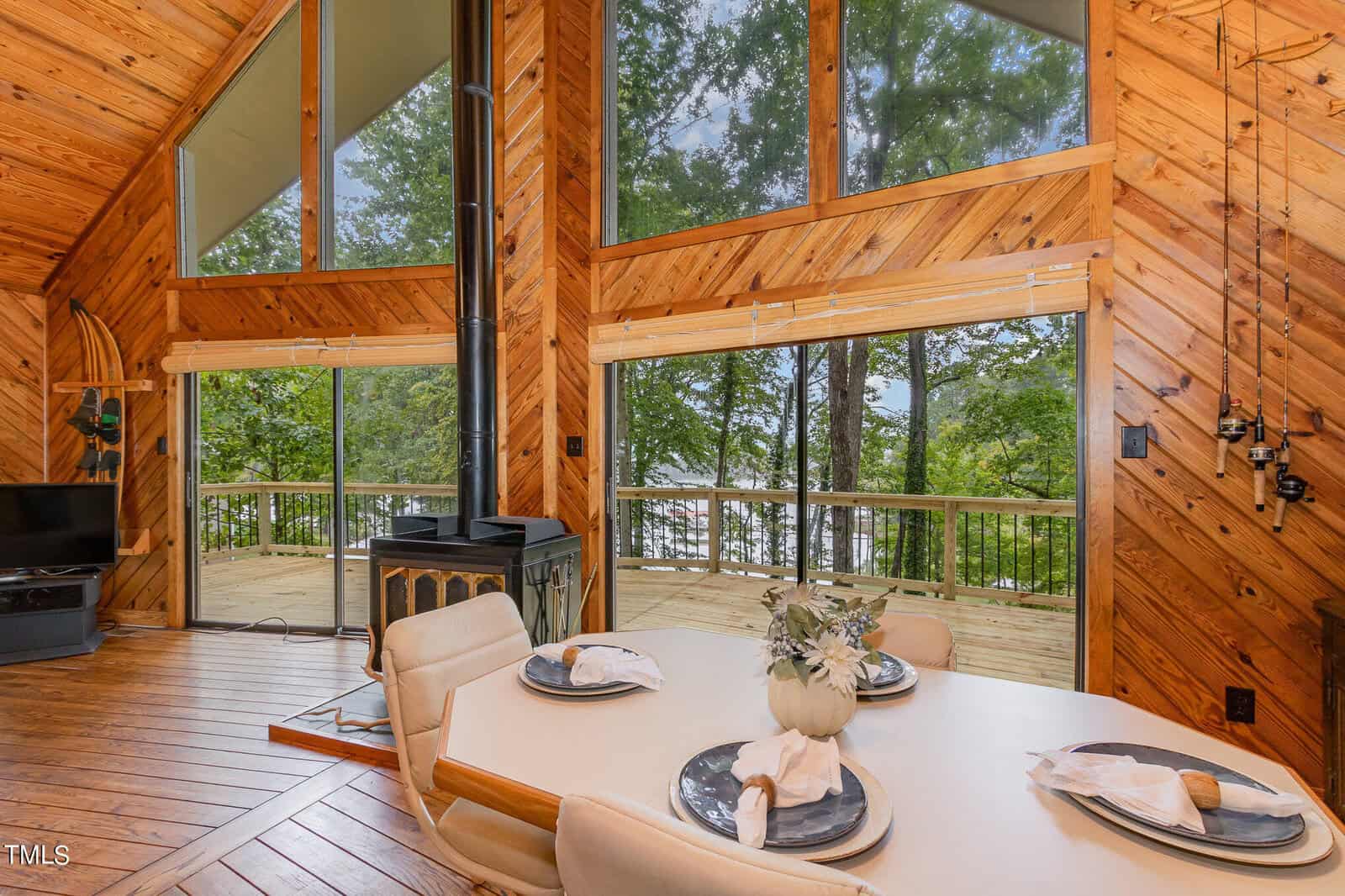 A cozy wooden cabin interior featuring a dining table with four place settings, a wood-burning stove, and large windows showcasing a scenic, tree-filled view with a spacious outdoor deck. The walls are paneled in knotty pine, giving a warm, rustic feel.