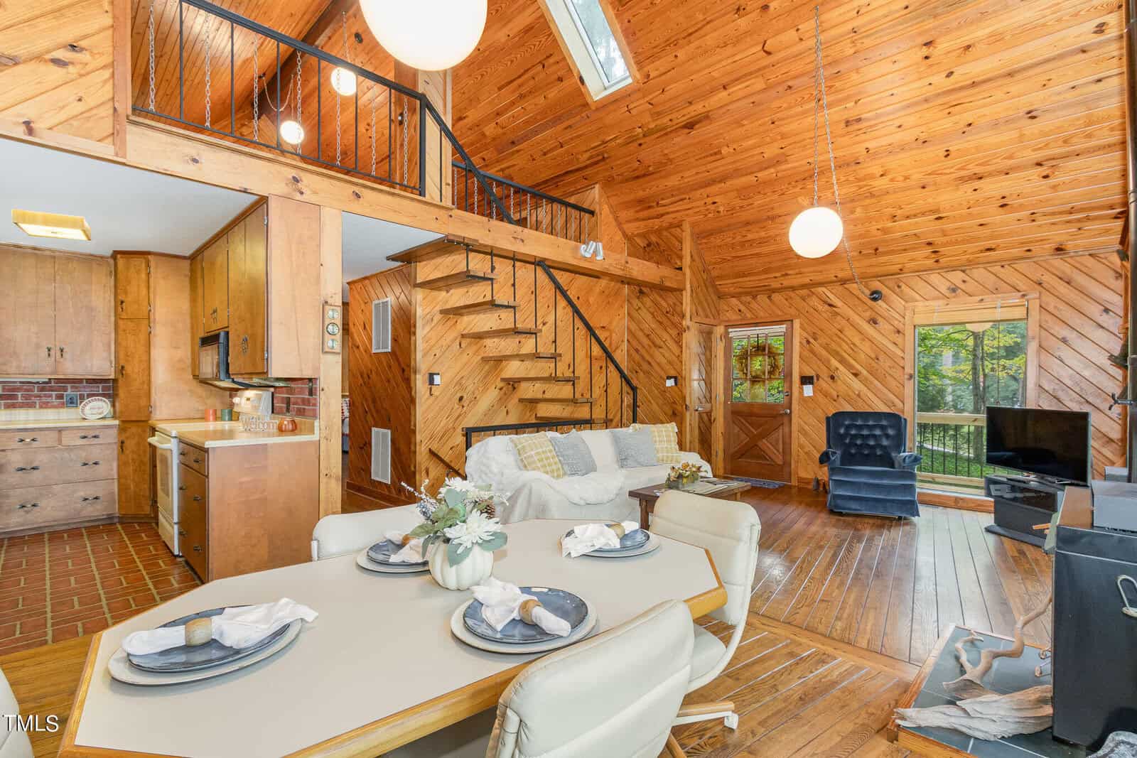 A cozy, wood-paneled interior of a cabin featuring a living space with a TV, blue armchair, and stairs leading to a loft. The dining area includes a table set for four. The kitchen has wooden cabinetry and brick accents. Skylights and large windows brighten the space.