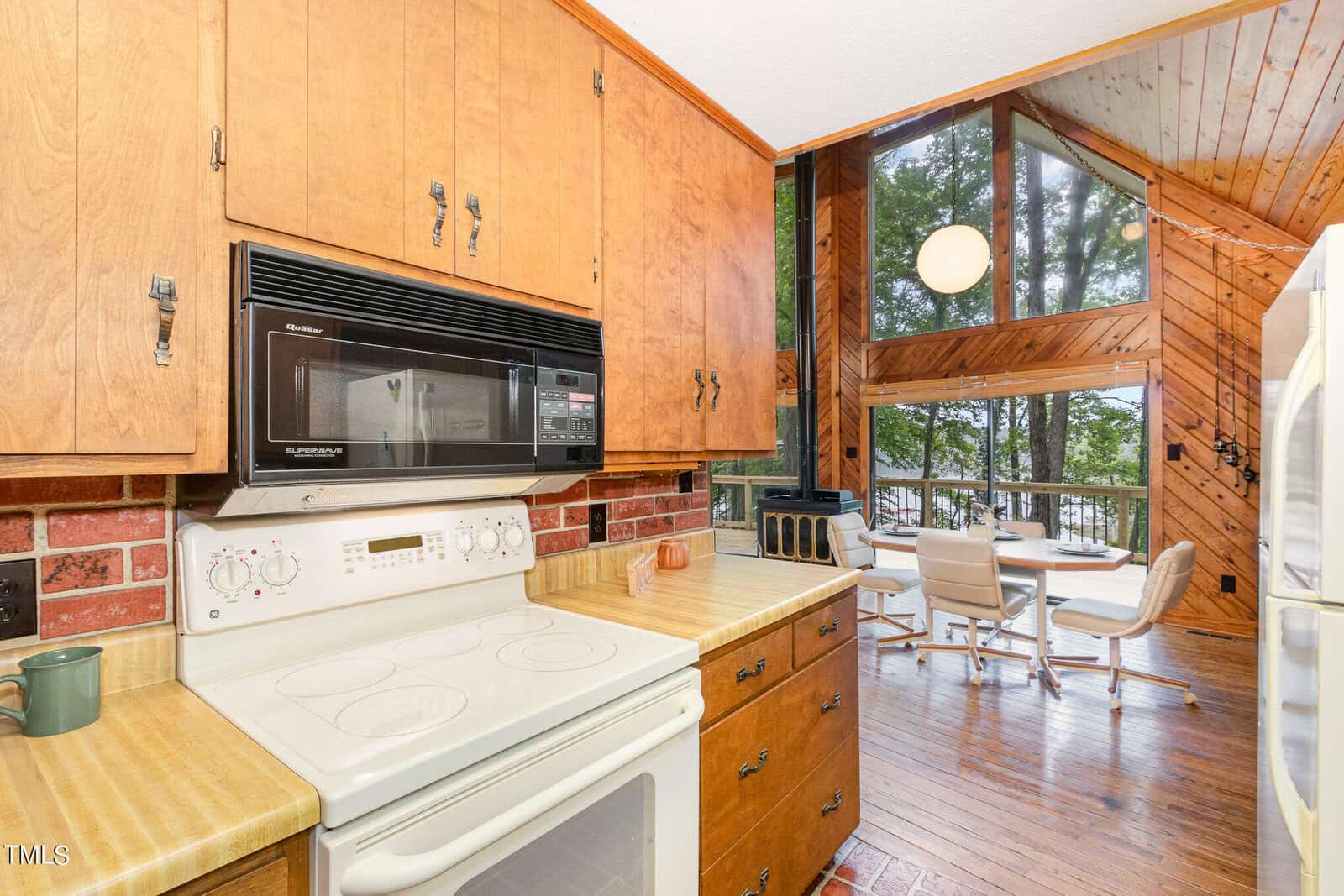 A kitchen with wooden cabinetry, white stove, and microwave. To the right is a dining area with a table, chairs, and a large window showcasing a wooded outdoor view. Wood paneling covers the walls and ceiling, adding a rustic touch.