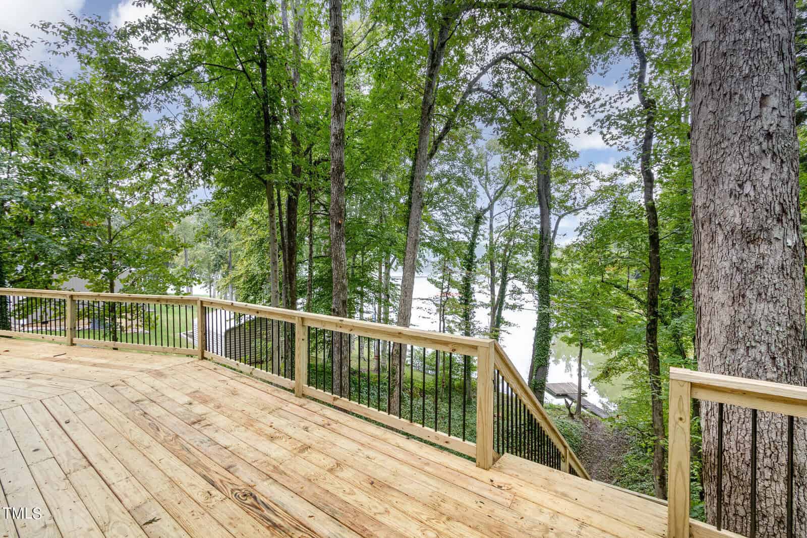 A wooden deck with a railing overlooks a serene lake surrounded by tall, dense trees. The lush green foliage and the calm water create a peaceful natural setting. The deck appears spacious and well-maintained, perfect for outdoor relaxation.