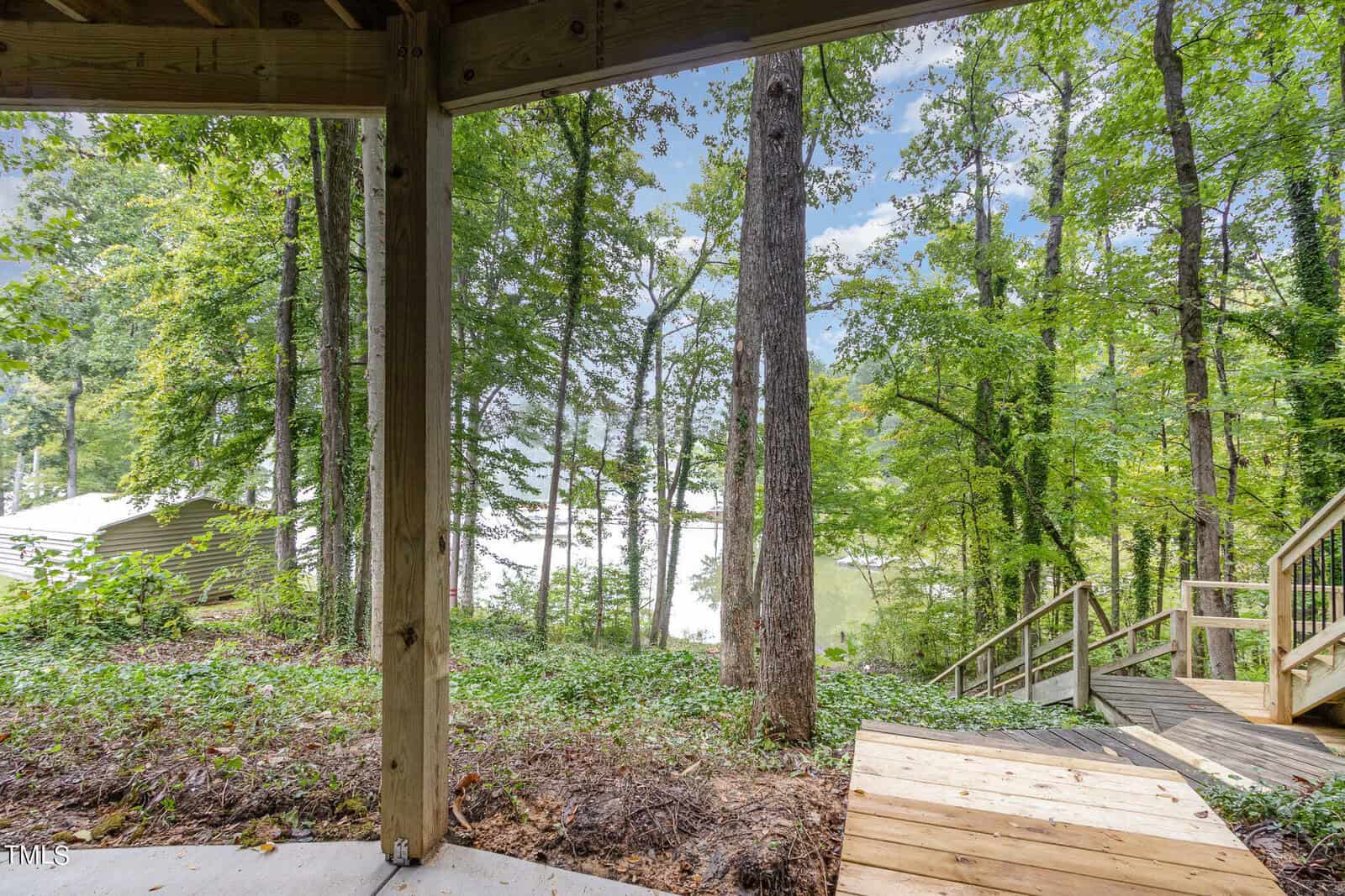View of a serene, wooded area with tall trees and a lake visible in the background. The foreground shows a wooden deck and stairs leading down to the water, partially shaded by the structure above. A house is visible on the left side of the image.