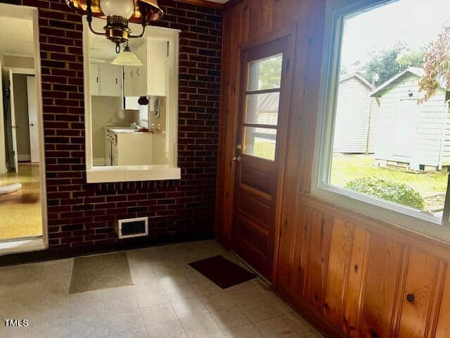A room features wood-paneled walls, a large window, and a wooden door leading outside. A brick wall with a small passthrough window opens to a kitchen area. A brass chandelier hangs from the ceiling. The floor has square tiles with a few brown rugs.
