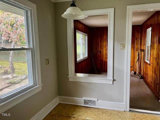 A small room with light gray walls and a window showing a tree outside. There's a wooden paneled opening leading to another room with wood-paneled walls. The floor has a light pattern, and a ceiling light fixture is visible.