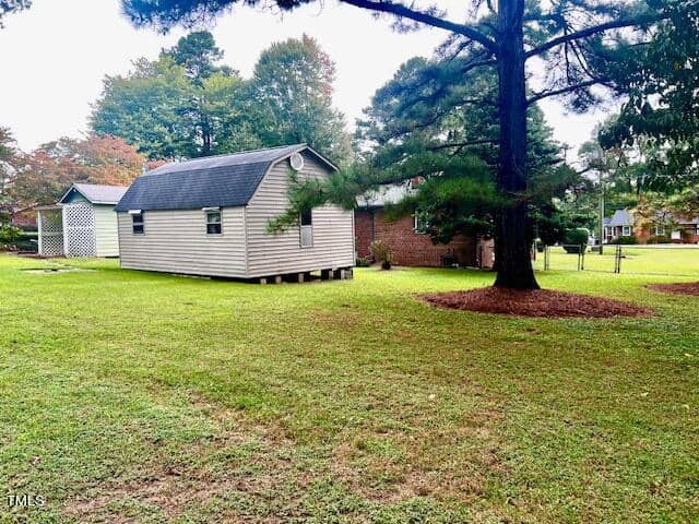A grassy backyard with a small beige shed on stilts. The yard has a large tree in the foreground and another small building and a brick house in the background. The sky is overcast.