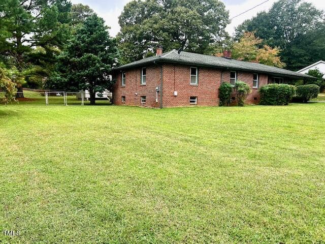 A brick house with a green roof sits on a large, well-maintained grassy lawn. The house is surrounded by tall trees, and the sky is overcast, creating a serene and peaceful atmosphere.