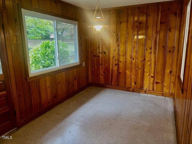 A small room with wooden panel walls and a large window overlooking greenery. The room has a hanging light fixture and a tiled floor. Natural light streams in through the window, creating a warm and inviting atmosphere.