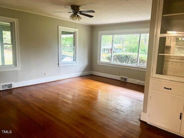 A spacious, empty room with polished wooden floors, large windows with views of greenery, and a ceiling fan. White walls and built-in shelving add to the bright, airy feel.