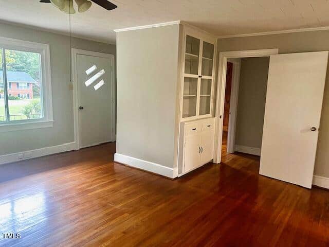 A bright, empty room with light gray walls and polished wooden floors. There's a ceiling fan, a window showing a partial view of a house, and a built-in white cabinet with glass doors. An open door leads to another room.