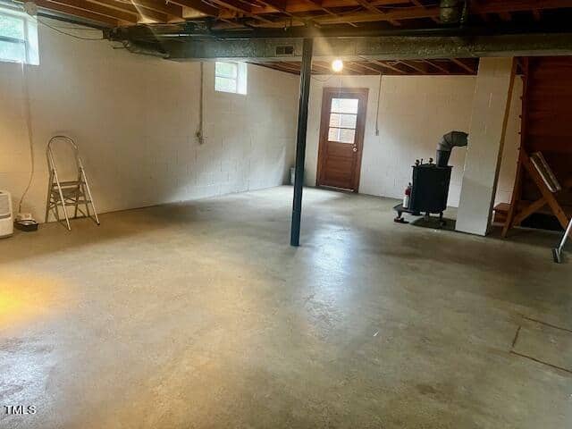 A clean, unfinished basement with concrete floors and cinder block walls. A wooden door with a window is illuminated by sunlight. A ladder stands against the wall, and a wood-burning stove with an exhaust pipe is situated to the right.