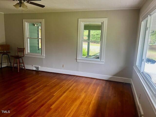 A bright, empty room with hardwood floors, light gray walls, and three windows. Two wooden chairs are placed in the corner near a ceiling fan. Outside, greenery and parts of a house are visible.