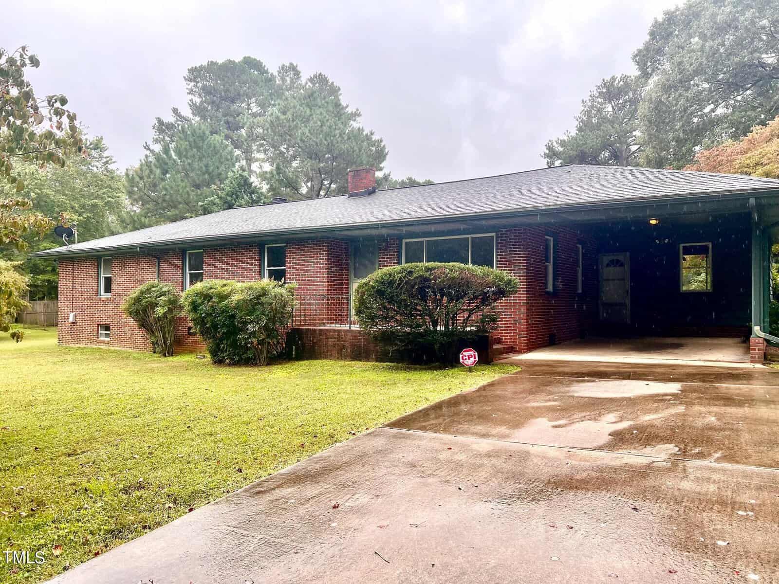 A charming brick single-story house at 718 Williamsboro Street, Oxford, features a gray roof and is surrounded by lush green shrubs and trees. A paved driveway leads to a carport on the right, glistening from the recent rain under the overcast sky.