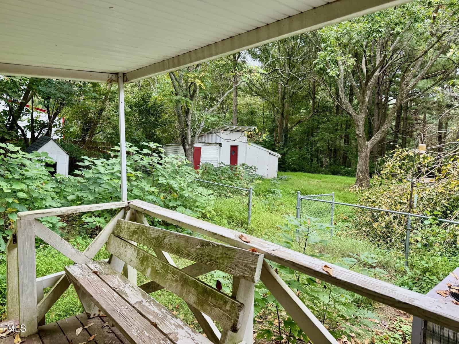 A backyard with a covered wooden porch featuring a weathered bench. The yard is lush with vegetation and trees, and a white storage shed with red doors is visible in the distance. A metal fence runs along the edge of the property.