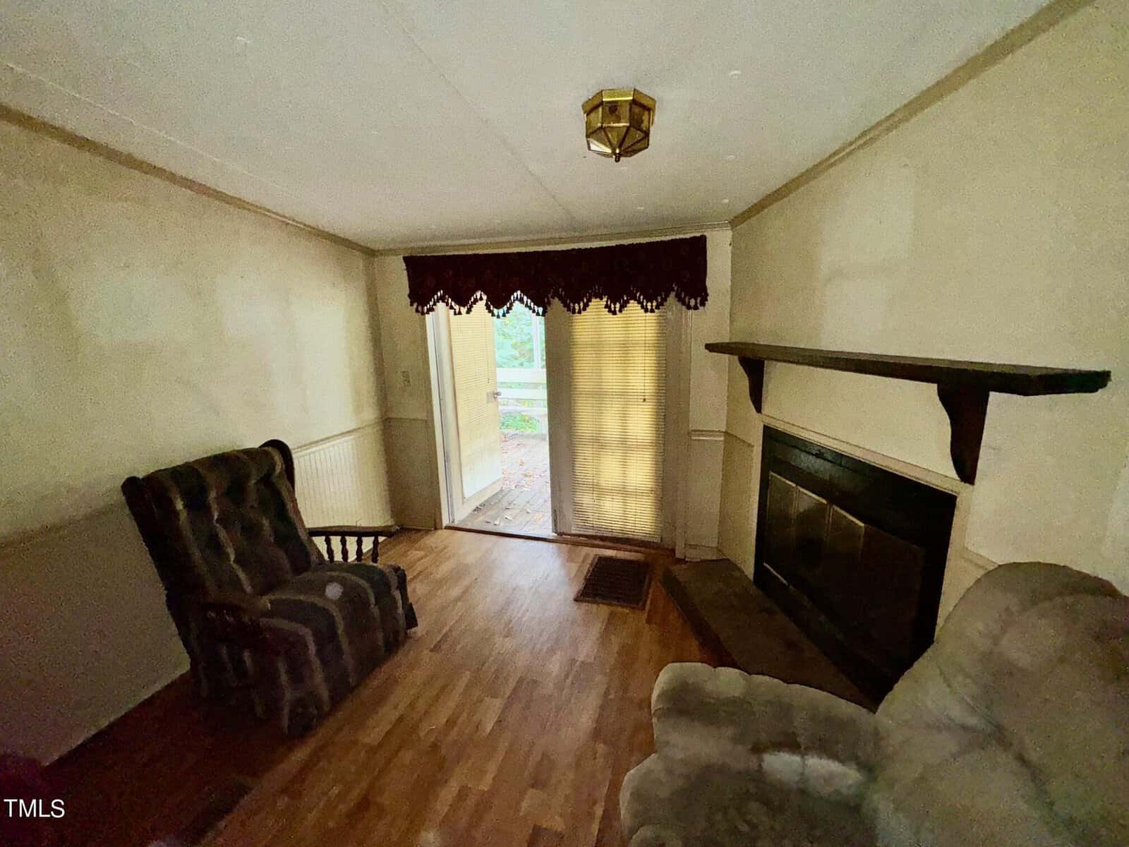 A modest living room with a dark wooden rocking chair and a cushioned armchair facing a fireplace. Light filters through glass doors covered by blinds and a valance, leading to an outdoor deck. The room has a wooden floor and plain, light-colored walls.