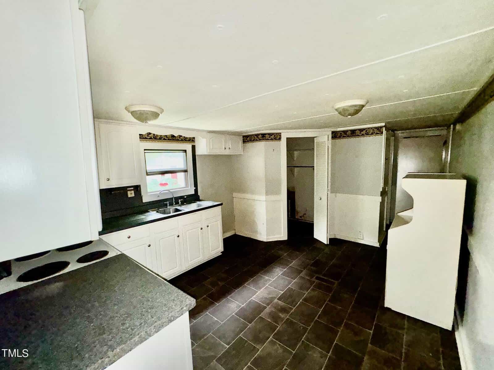 A kitchen with white cabinets, dark countertops, and a small sink under a window. The room has dark tile flooring and a stove on the left. There is a small open closet and a doorway to the right. The walls are light-colored with a decorative border.