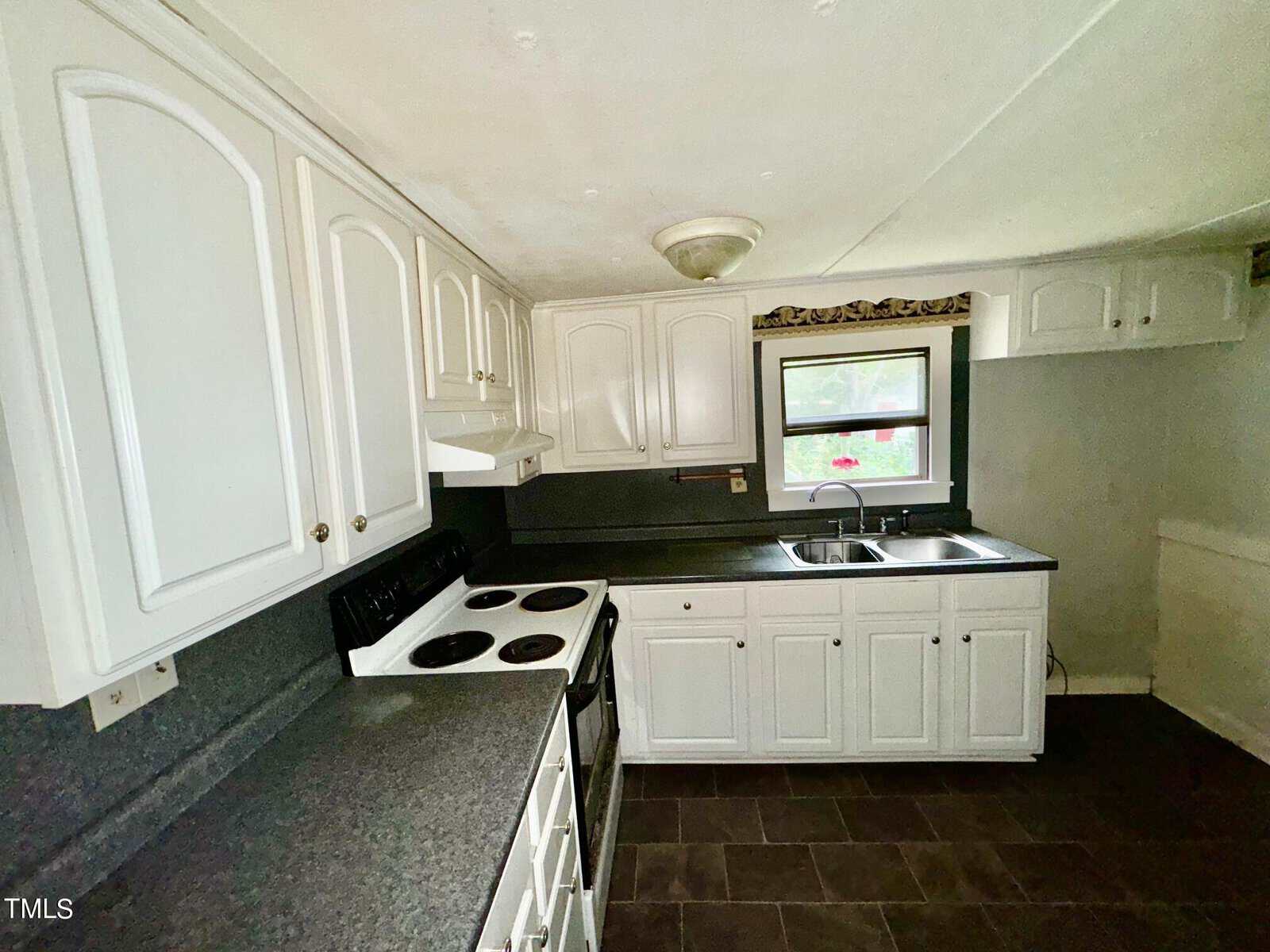 A small kitchen with white cabinets, a black countertop, and a black electric stove. The floor is dark tile. A window above the double sink lets in natural light. A ceiling light fixture is visible.