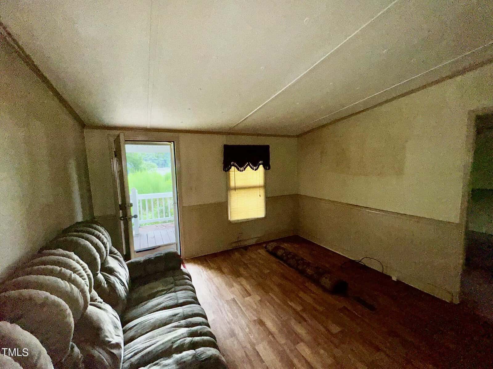 A dimly lit room with light-colored walls, a wooden floor, and a green, fabric sectional sofa. An open door leads to a deck outside. A window with a black valance is centered on the back wall, allowing some natural light in. The room is mostly empty and appears to be in need of some finishing.