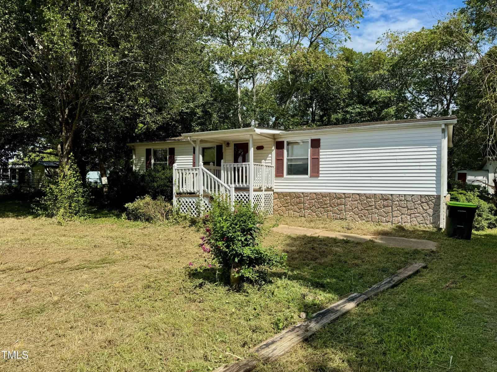 A small white house with a stone foundation, located at 1611 Phyllis Lane, Henderson. Surrounded by trees and a grassy yard, it features a covered porch with railings and a path leading to the entrance. A trash bin is visible to the right.