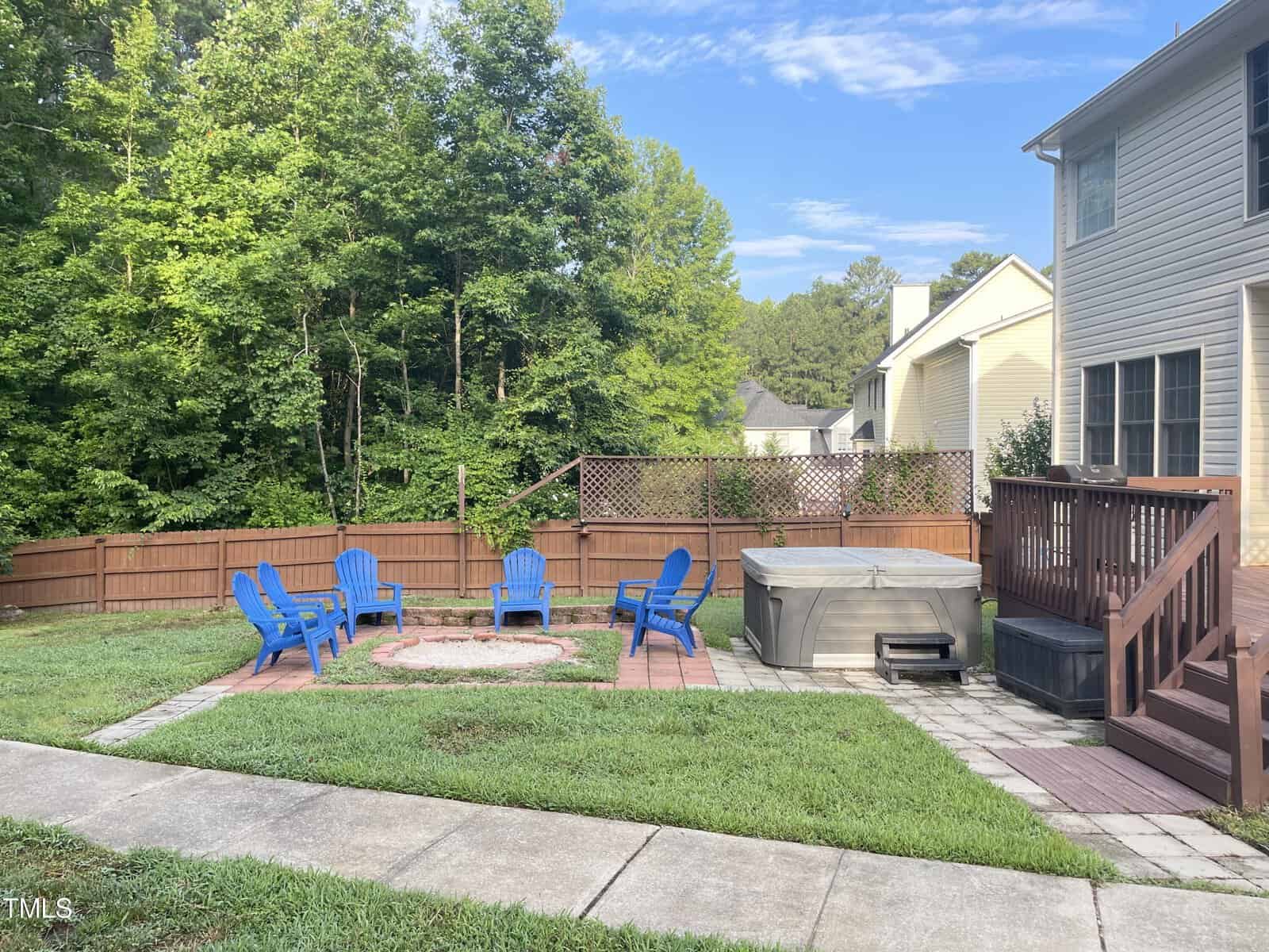 A backyard with a fire pit surrounded by six blue chairs on a grass patio. A hot tub is adjacent, near wooden stairs leading to a house. The area is enclosed by a wooden fence and bordered by trees and another house in the background.