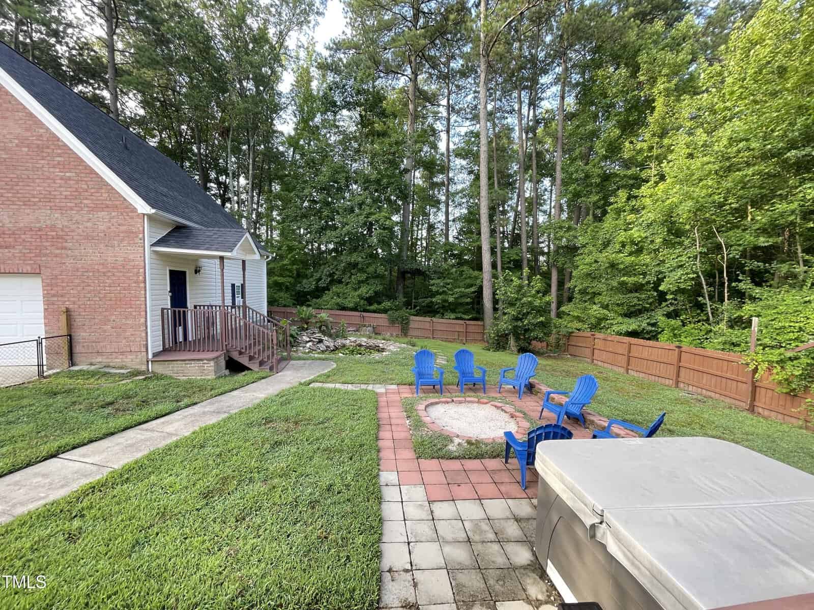 A backyard with a brick house and a square patio featuring a fire pit surrounded by blue chairs. A hot tub is in the foreground, and tall trees and a wooden fence line the background.