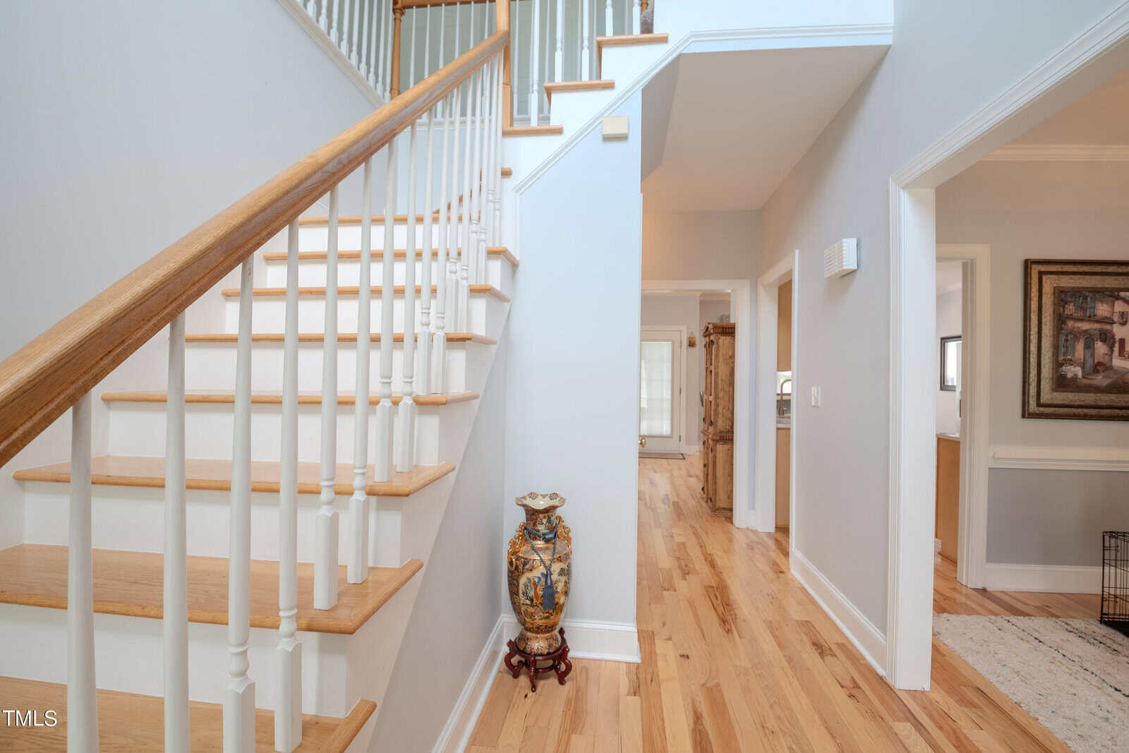 A bright hallway with light wood flooring and white walls features a wooden staircase with white balusters on the left. A decorative vase is placed at the base of the stairs. An open doorway leads to a room with framed artwork on the wall.