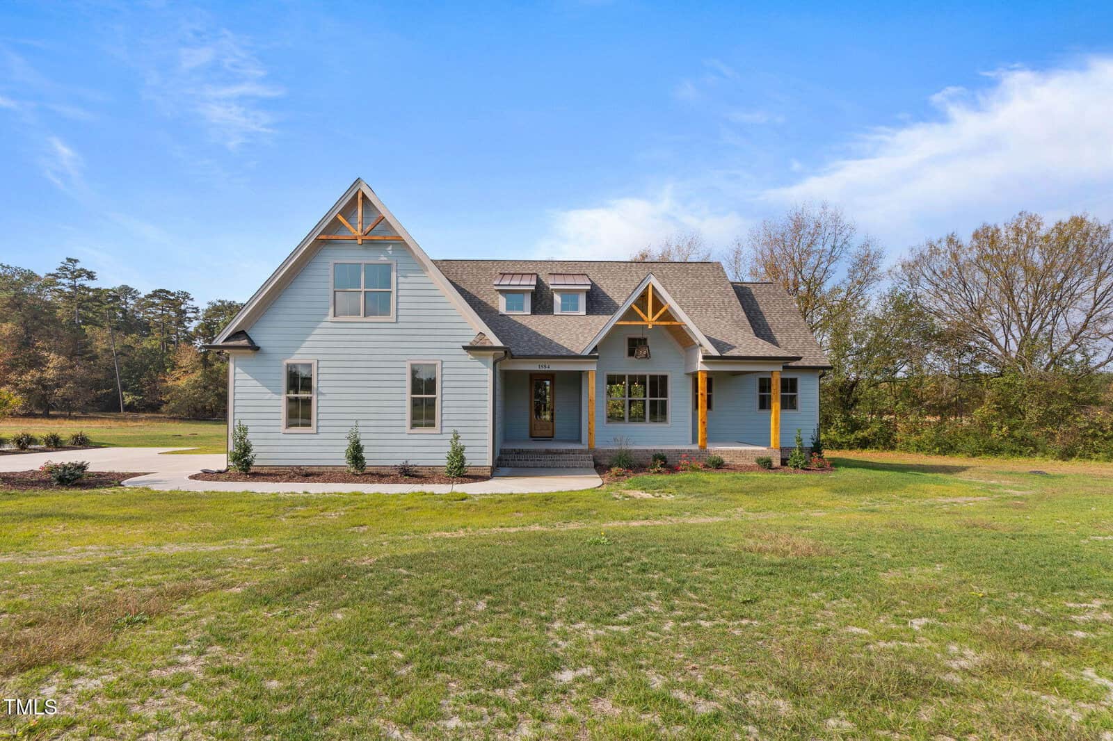 A light blue two-story house with a gray roof, wooden accents, and a covered front porch, set on a grassy lawn with trees in the background under a blue sky with scattered clouds.