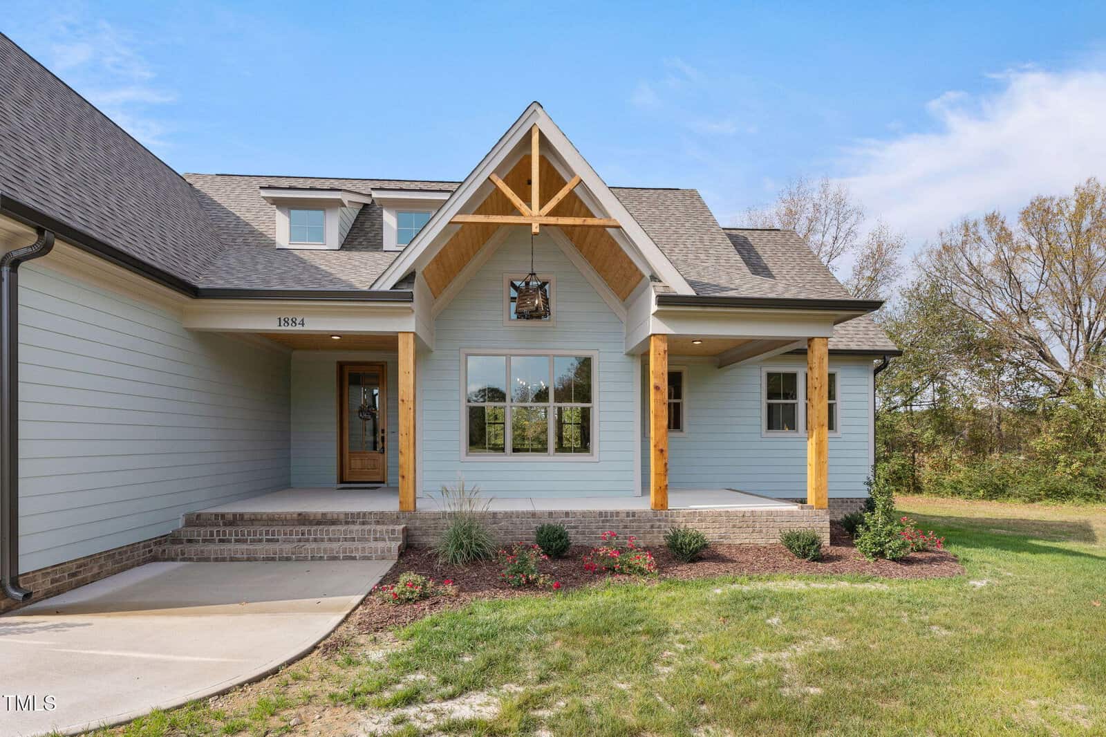A modern house with light blue siding, wooden posts, a covered front porch, brick steps, and a gabled roof. There are large windows, a wooden front door, minimal landscaping, and a grassy yard.