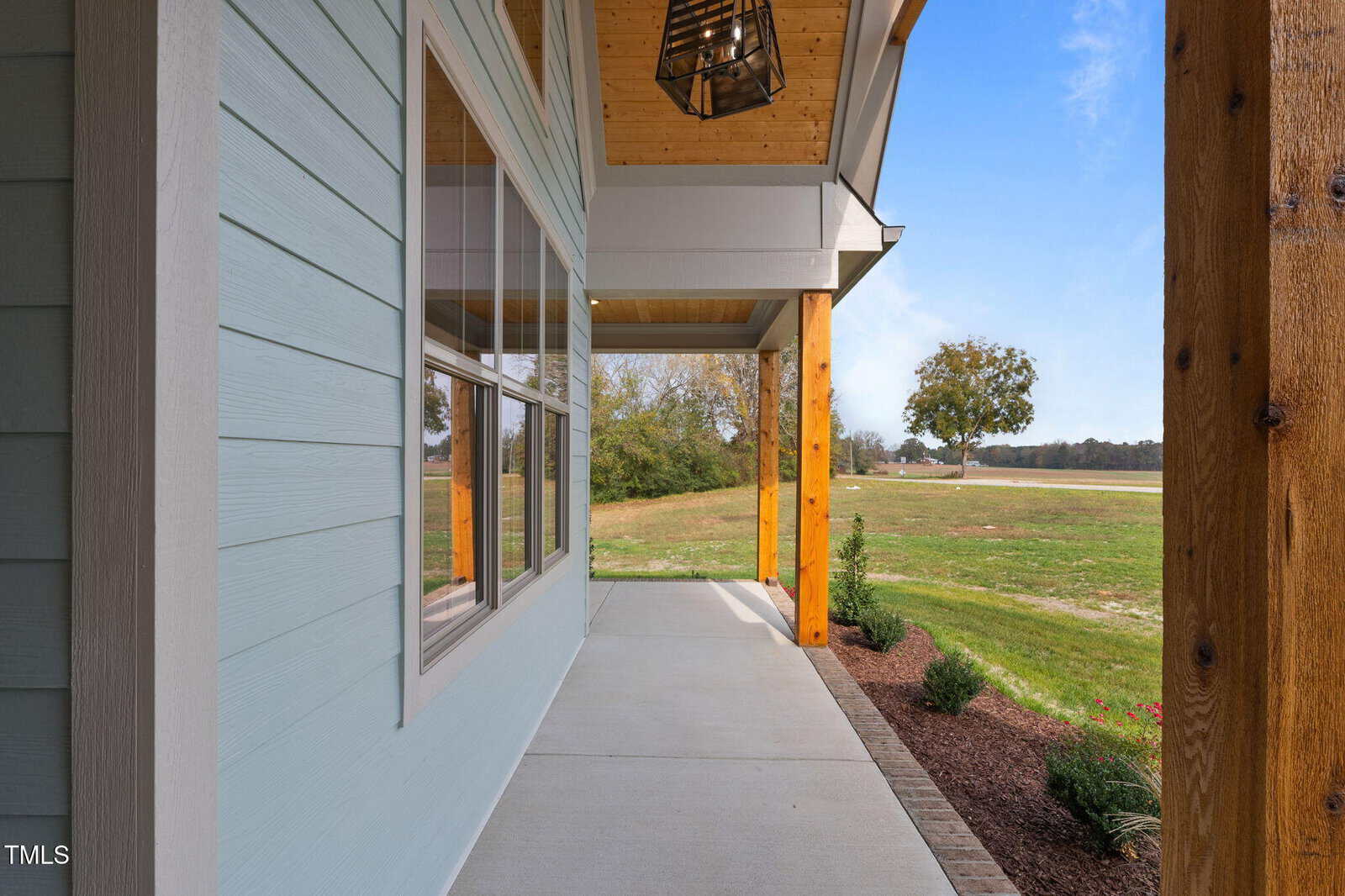 A covered front porch with light blue siding, large windows, wooden columns, a hanging light fixture, and a view of a grassy yard with a tree in the distance.