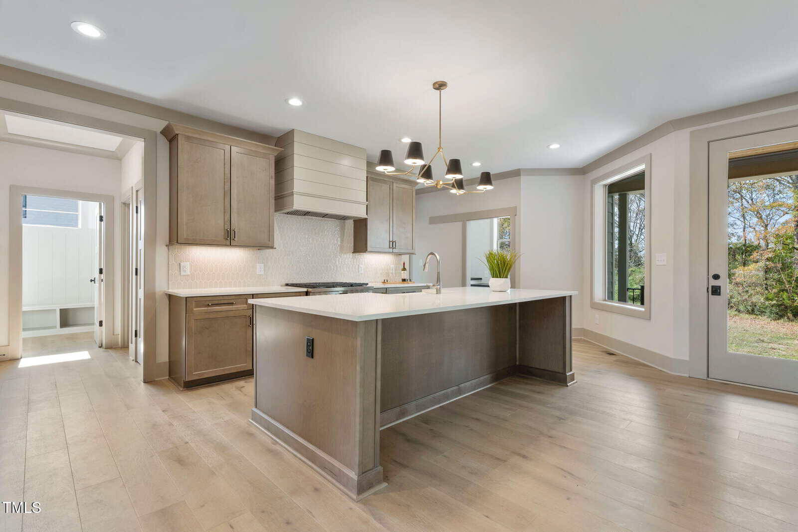 A modern kitchen with light wood floors, taupe cabinets, a large island with a sink, a black and gold chandelier, and large windows letting in natural light. A door leads outside to a grassy yard.