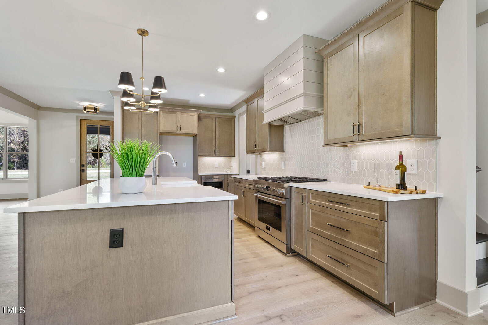 Modern kitchen with light wood cabinets, white countertops, a central island with a plant and sink, stainless steel appliances, a wine bottle on a tray, and geometric backsplash tiles. Recessed and pendant lighting overhead.
