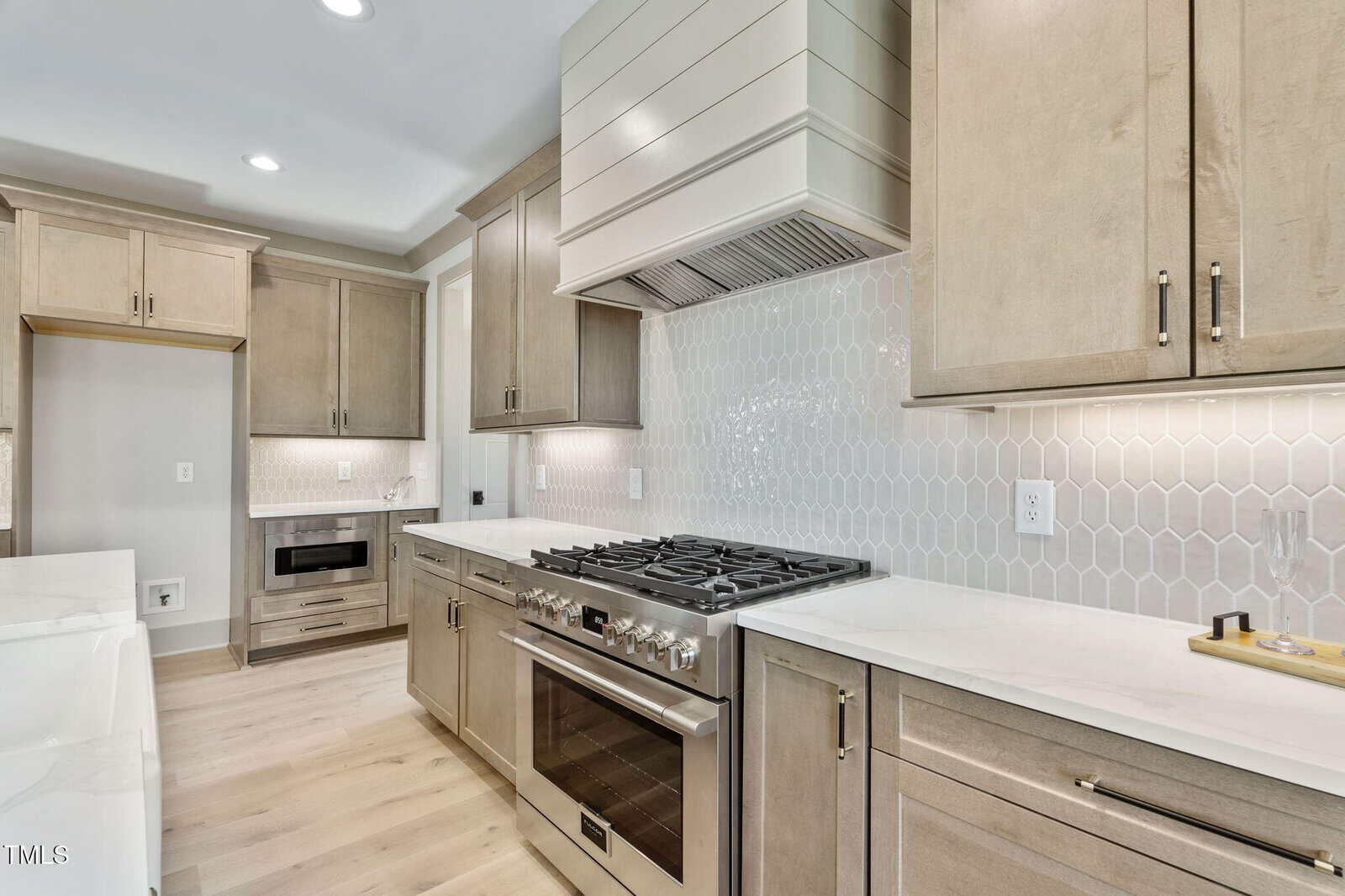 A modern kitchen with light wood cabinets, a stainless steel gas range and oven, white countertops, a white farmhouse sink, and a backsplash with white hexagonal tiles. The area is well-lit with recessed lighting.