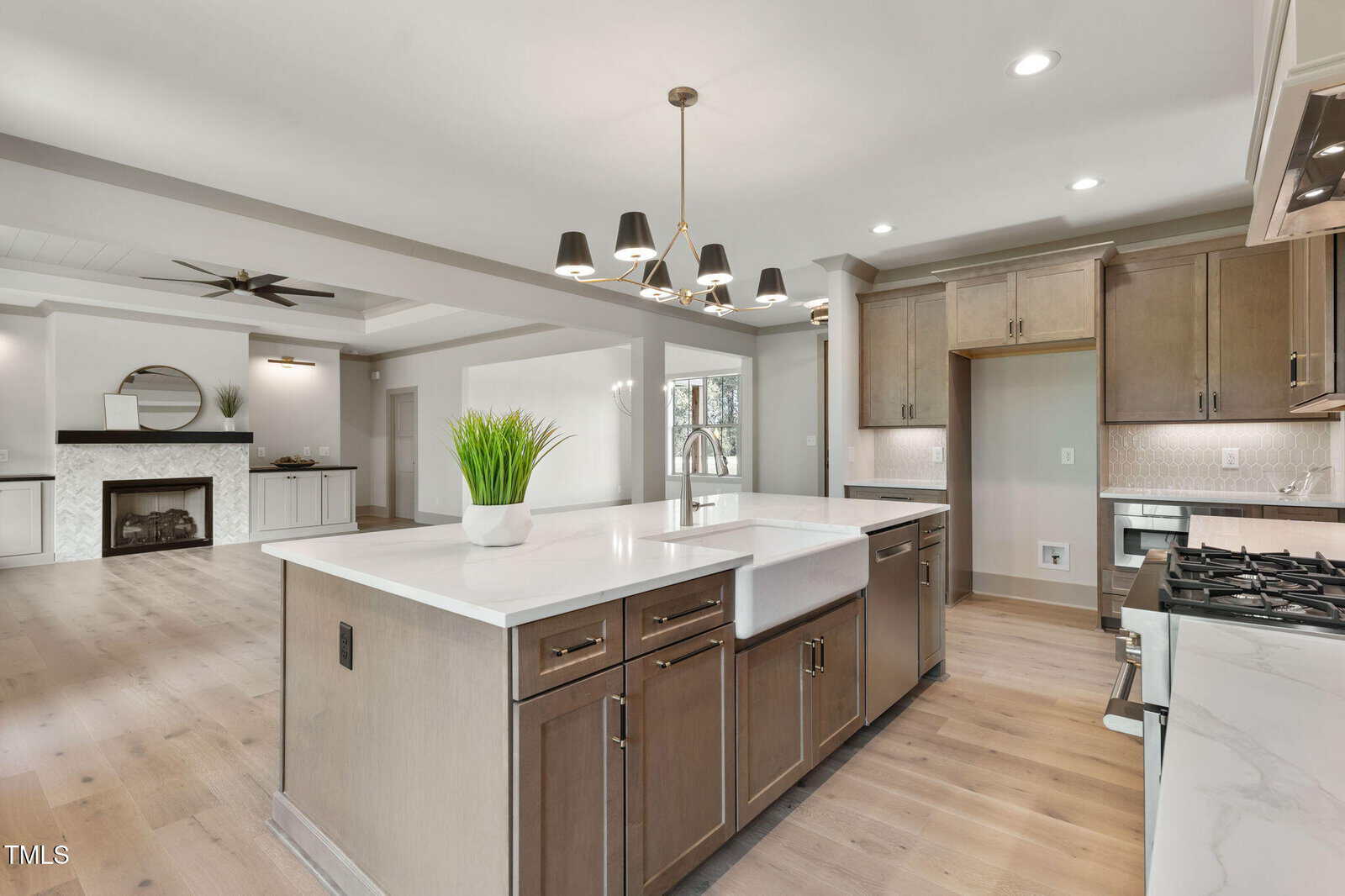 A modern kitchen with light wood flooring, a large island with a white countertop and farmhouse sink, stainless steel appliances, light brown cabinets, and a dining chandelier. A living area with a fireplace is visible in the background.